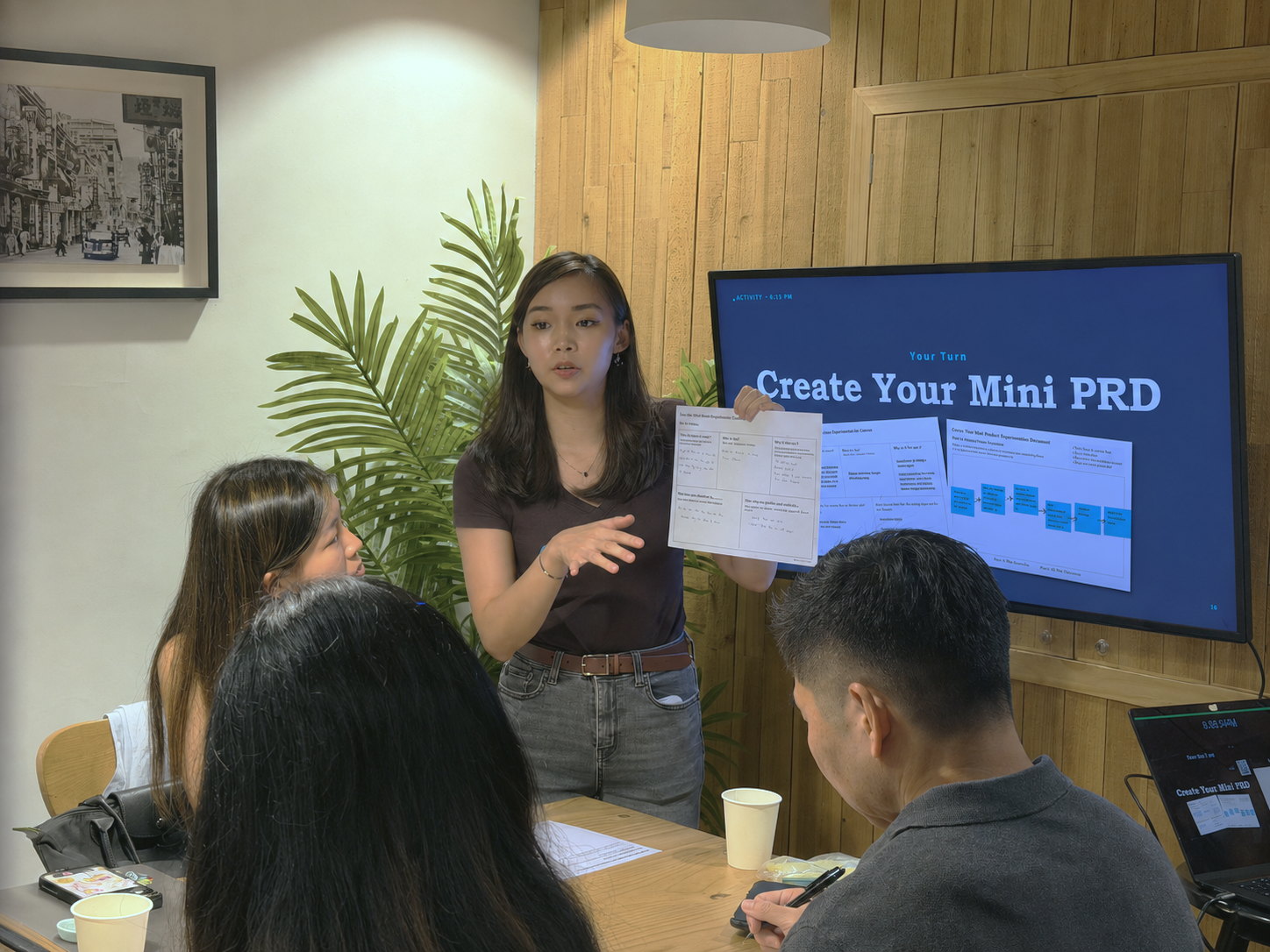 Sharon standing mid-sentence, holding up a completed worksheet to a small group seated at a table.