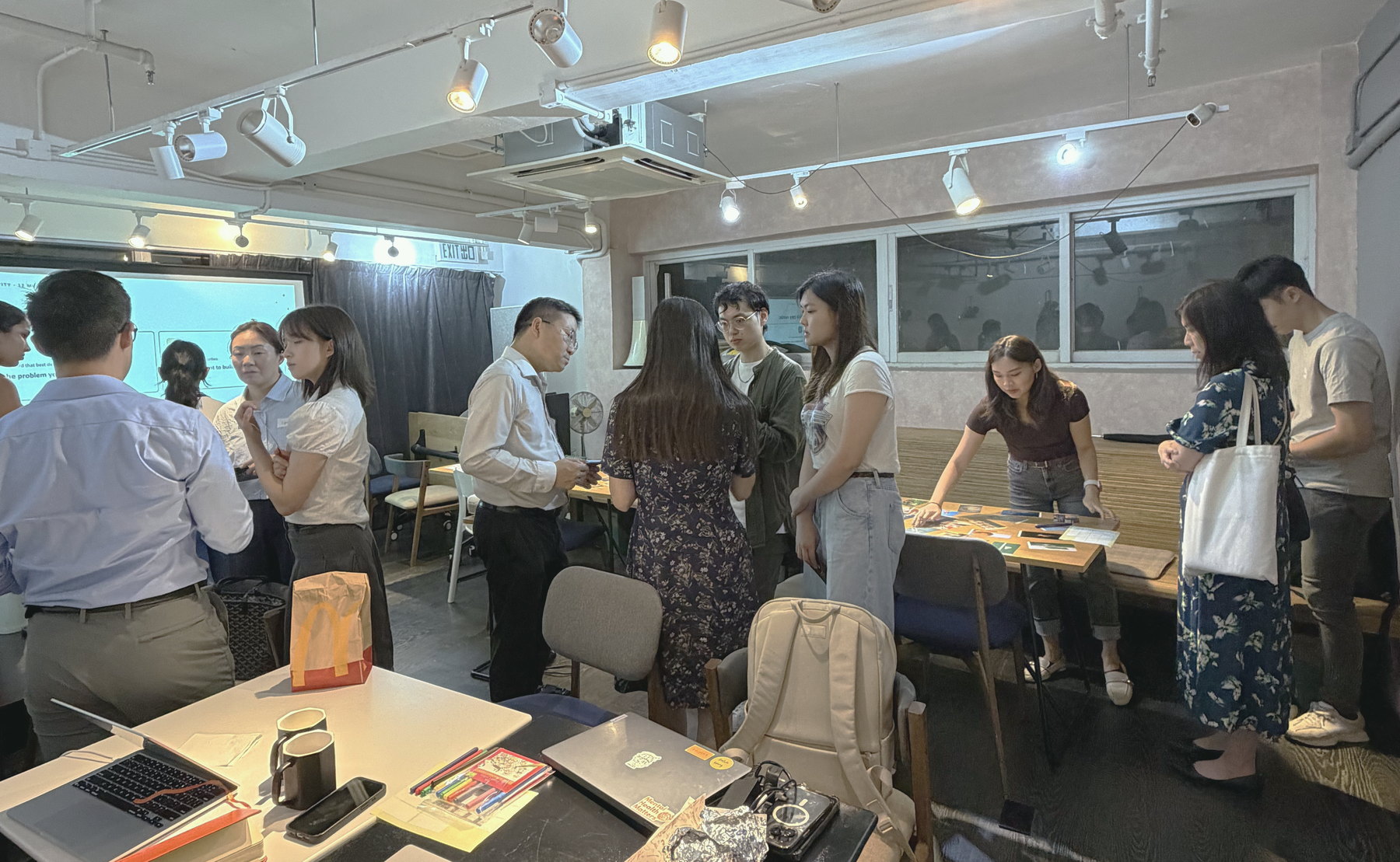 Participants standing and talking in groups around tables at the start of a session, bags and coffee cups still out.