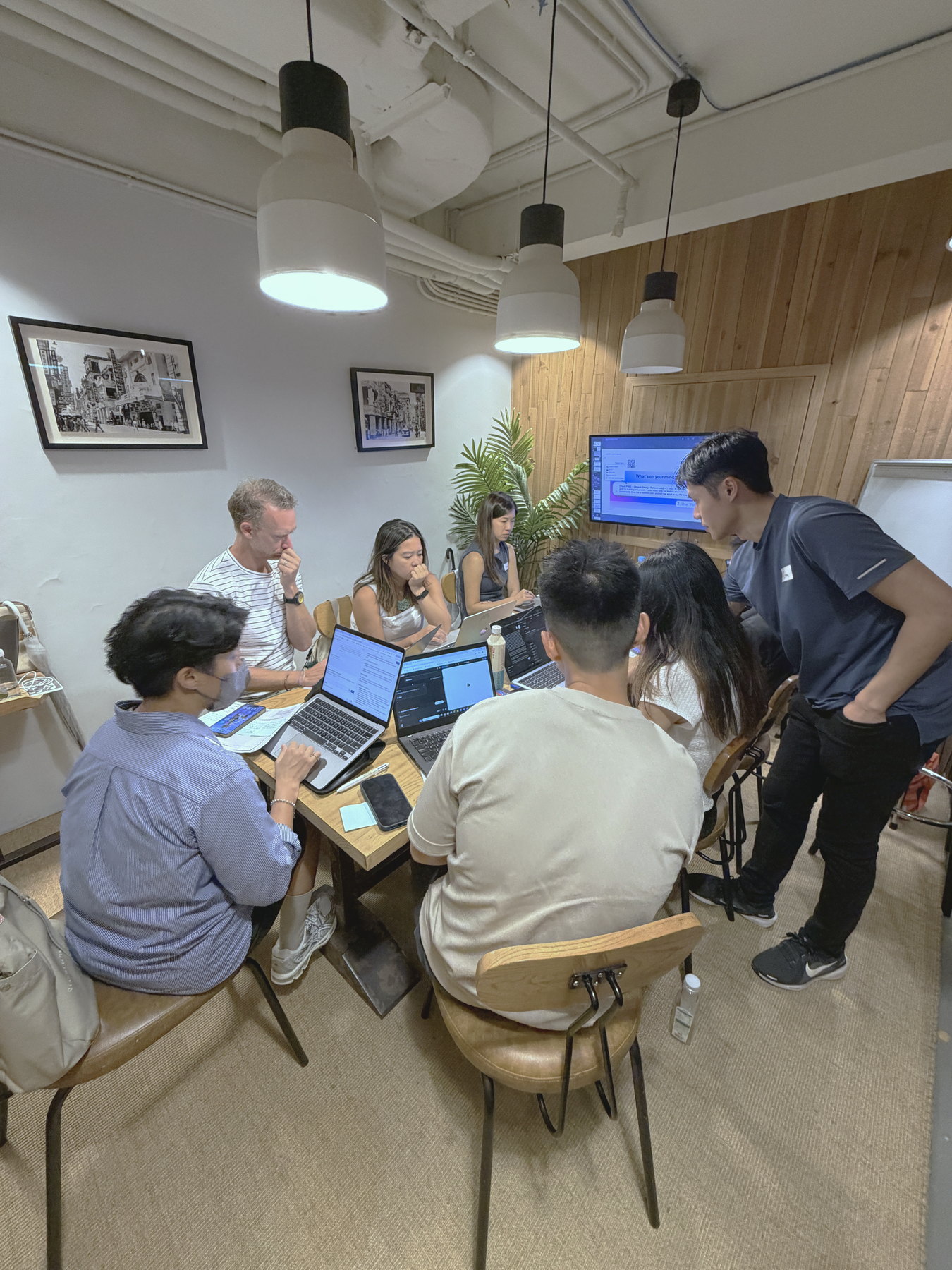 Participants gathered around one table with laptops open, one person standing to help another with what is on screen.