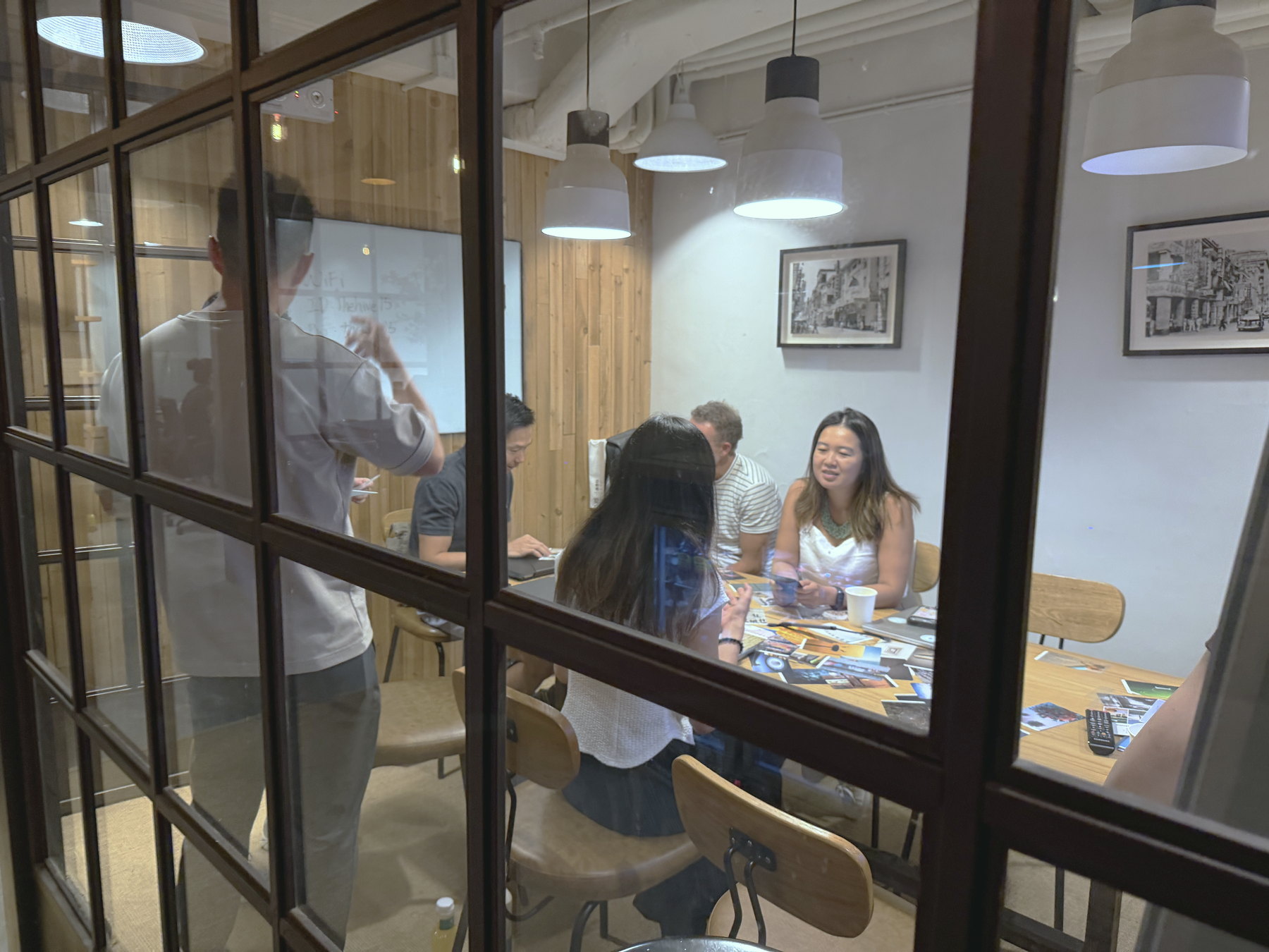 The workshop seen from outside through a glass partition, participants talking around a table under pendant lights.