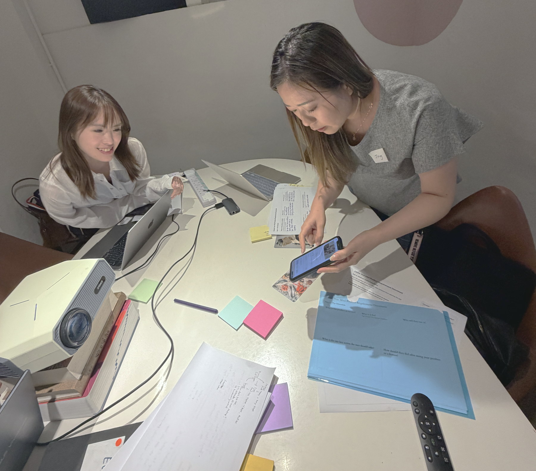 A participant holding up her phone to show the app she built, another participant leaning in to look, the table between them covered in notes and sticky notes.