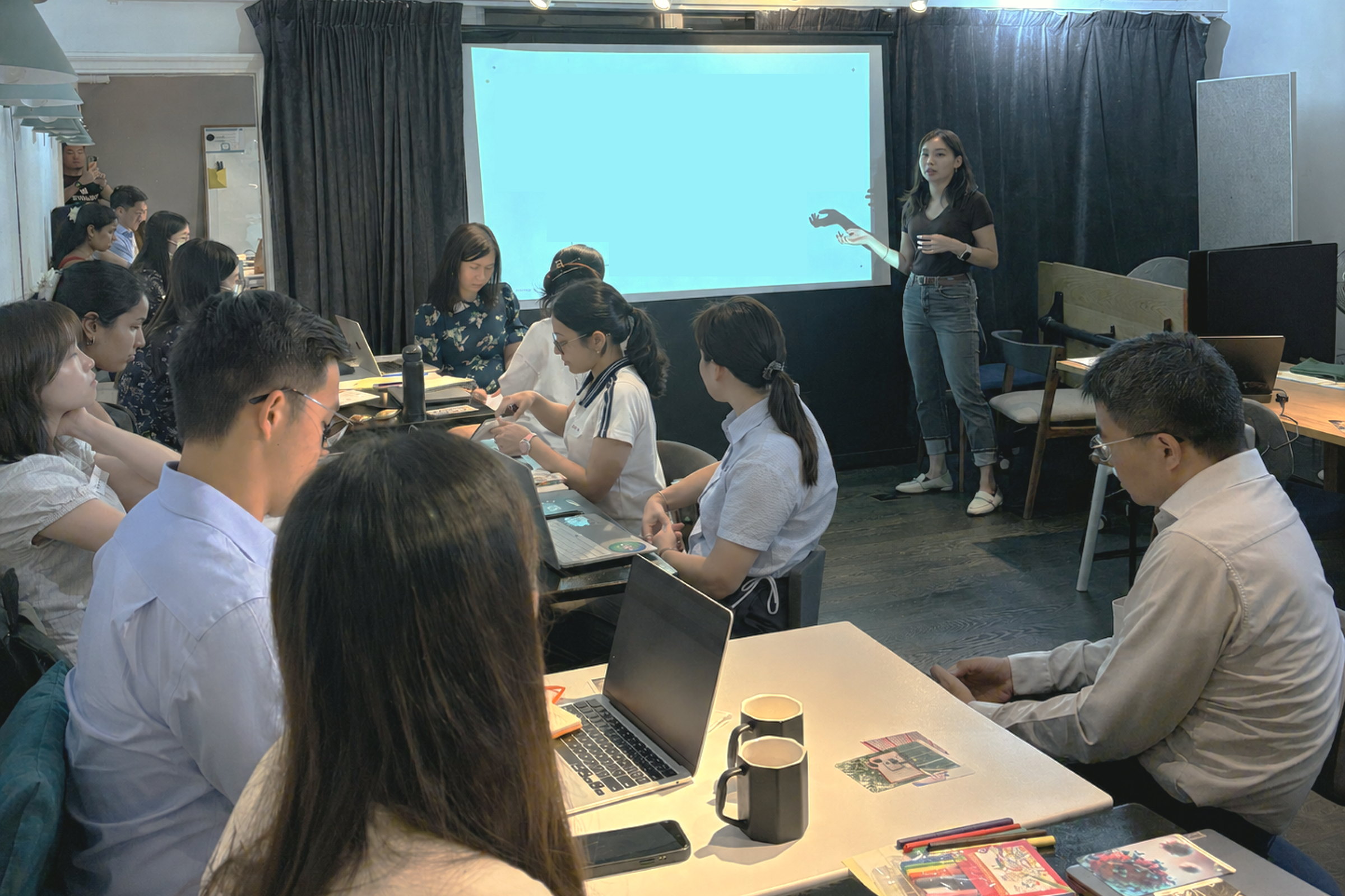 Sharon presenting to a full room of seated participants at a Mind Made workshop, laptops and printed worksheets on the tables in front of them.